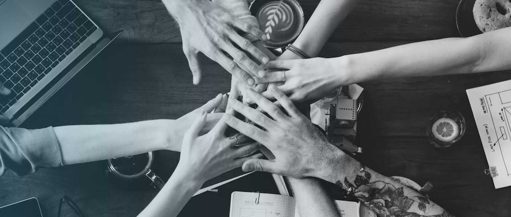 Greyscale image of hands-in/team huddle over an office desktop